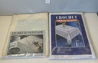 Two packaged tablecloths: the Quaker-Mayfair brand oval natural cotton-polyester cloth on the left, and a white rectangular vinyl crochet tablecloth on the right