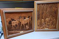 Photo showing two framed wood carved panels side by side, one depicting 'Mother and Colt' with horses and dog, the other 'Maple Syrup' with a horse and sap collection scene.