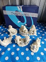 Six Snow Babies figurines displayed on a blue polka dot surface with a labeled box behind them. Various poses depicting children and snow-related themes.