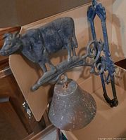 Photo showing the cast metal doorbell featuring a detailed cow figure on top, conical bell, and decorative mounting bracket with visible aged patina and surface wear.