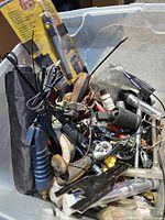 Various miscellaneous tools and items inside a clear plastic bin including an engraver tool with a blue handle and several cables, a flashlight, wrenches, and other hardware.