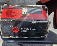 Photo of the original box showing the Mac Tools logo and product specifications including 2 inch angle grinder, 20,000 RPM, built-in comfort grip, safety lever, side exhaust, included accessories, and grit wheels.