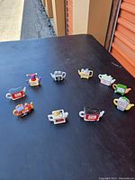 Photo shows nine small collectible ceramic teapots mostly white or cream with a red rose design. Teapots are in various shapes and color accents, including two red teapots with paint peeling.