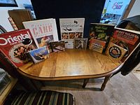Seven cookbooks and five music CDs arranged on a wooden table, showing the variety in cuisine and music genres.