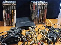 Sony PlayStation 2 console with 12 PS2 game cases arranged upright, two black controllers with cables, composite AV cables, and power adapter on a wooden table with dark background.