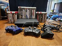 Photo showing the PlayStation 3 console, three wired controllers (one blue, two black), and 36 game cases arranged behind the console.