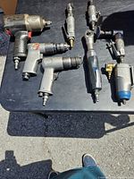Top view of eight different air tools arranged on a black table. Visible details include air hose connectors, pistol grip handles, and metal bodies of tools.