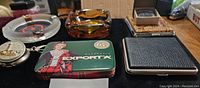 Photo showing Exporta tin box, two ashtrays (amber glass and ship's wheel shape), keychain, and lighter box