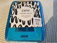 Blue DSW portable art kit case with label indicating contents: 10 sheets of white paper, 8 mini washable markers, and 32 crayons. Front plastic shows partial tear but sealed.