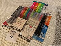 Top view of unopened marker, pen, and highlighter packs arranged on beige fabric surface.