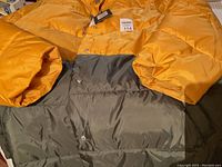 Photo showing men's winter coat with mustard yellow top half and sleeves and dark gray bottom half. Coat is puffy and insulated with snap button closure visible.