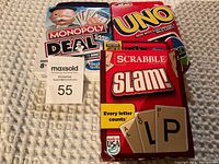 Photo showing three boxed card games Monopoly Deal, Uno, and Scrabble Slam! stacked on carpet.