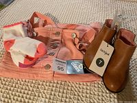 Pink denim dress, brown ankle boots with tags, and three pairs of white socks with pink/red accents displayed on a textured beige surface.