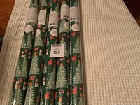 Five rolls of Christmas wrapping paper with festive tree and gift design laid on a white blanket, showing the tops and full length of the rolls.