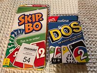 Photo showing Skip-Bo and DOS card game boxes side by side. Skip-Bo box is white with colorful cards pictured and DOS box is blue with large game name and various cards shown.