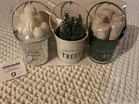 Front view of the set of three Christmas-themed metal buckets sealed in original packaging on a textured cream blanket.