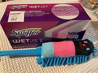 Box of 15 Swiffer Wet Jet Pads beside a colorful cat-shaped microfiber duster with a blue plastic handle on a beige textured surface.