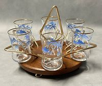 Front view of six shot glasses in circular brass and cherry wood caddy, showing floral pattern and gold rims on glasses.