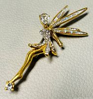 Close-up photo showing the brooch in detail, featuring the gold-tone fairy with head, body, and wings embellished with clear crystals on textured white background.