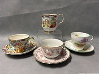 Three vintage porcelain teacup and saucer sets pictured on a gray background including Royal Albert Old Country Roses mug, pink floral demitasse set with spoon, and multicolor floral set.