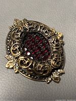 Close-up photo of the brooch showing the ornate filigree metalwork and red crystals in the center against a neutral background.