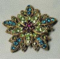 Close-up of the gold-tone flower brooch featuring blue, green, and purple crystals set in petal shapes on a textured pale surface.