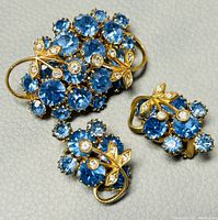 Set of vintage gold-tone brooch and earrings on light textured surface showing blue and clear crystals with leaf accents.