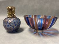 Fragrance lamp with brass top and speckled blue glass body next to blue, pink and gold striped pedestal bowl