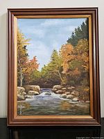 Full view of framed oil painting showing autumn river scene with rocks and trees.