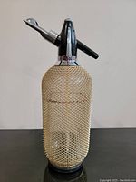Full view of vintage soda siphon bottle with metal mesh covering, black top, and chrome accents