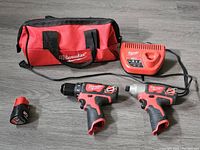Photo of two Milwaukee M12 cordless drills, one battery, charger and red contractor bag on wooden floor.