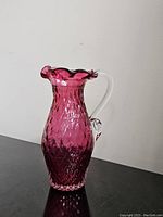 Front angled view of the cranberry diamond cut art glass pitcher with a clear handle and ruffled rim, standing on a dark surface against a white wall.