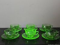 Six green uranium glass cups with matching saucers arranged in two rows on a dark surface. Cups are translucent and glowing under UV light.
