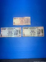 Three Indian banknotes placed on a blue surface: one 10 rupee note and two 100 rupee notes.