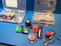 Top view of two plastic tackle boxes with various fishing lures and soft baits, plus a fishing reel and assorted fishing accessories on a blue surface