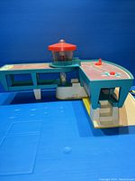 Front and side perspective of vintage Fisher-Price airport playset showing control tower with figurines inside, runway surface with printed markings, and stairs to elevated section.