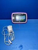 Front view of LeapPad 3 tablet displaying screen and connected charger, lying on blue surface with stylus attached.