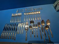 Full view of all flatware pieces laid out on a blue surface showing knives, forks, spoons, and serving utensils.