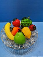 Vintage glass fruits including banana, apple, orange, pear, lemon, grapes, and another red fruit displayed in a clear crystal bowl with cut glass design on blue background.