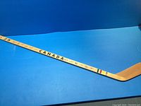 Full length view of the wooden hockey stick showing natural wood finish with 'CANADA' printed in black on the shaft, traditional Hudson Bay stripes near the blade end, set against a blue background.