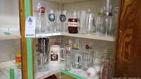 Shelf with various beer glasses and mugs featuring logos from breweries like Moose Brew, Sapporo, and Heidilberg. Includes clear, frosted, and colored glasses and ceramic steins.