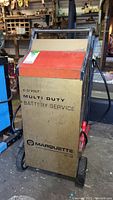 Front view of Marquette battery service unit showing brand name, voltage range and multi duty label with attached clamps on the front sides.