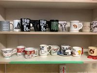 Wide view of two shelves containing 16 assorted branded mugs including novelty gun mugs, Disney characters, Campbell's soup mugs, and aviation-themed items.
