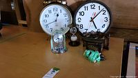 Photo showing multiple clocks including a large round white wall clock, gold twin bell alarm clock, small glass dome clock with rotating pendulum, hourglass sand timer, black decorative cuckoo clock, and green cloth item on wooden surface.