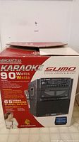 Photo of the karaoke machine packaging showing front and side views and listing features including 90 watts output and 65 songs included.