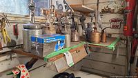 Photo showing eight metal sprayers and oil cans arranged on a bench with some background tools and signs.