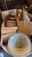 View of two vintage hanging light fixtures with ornate metal tops and frosted glass shades along with one round ceiling light shade in a cardboard box.