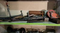 View of one shelf containing various vintage wooden handle and metal tools including hammers, hand planes, saws, and clamps.