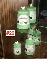 Three green and white pottery canisters with painted hens and rooster figurines on lids displayed on a glass shelf.