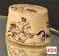 Front and side view of small hand decorated box showing stylized black illustrations of human figure riding animal and calligraphic markings on cream surface.